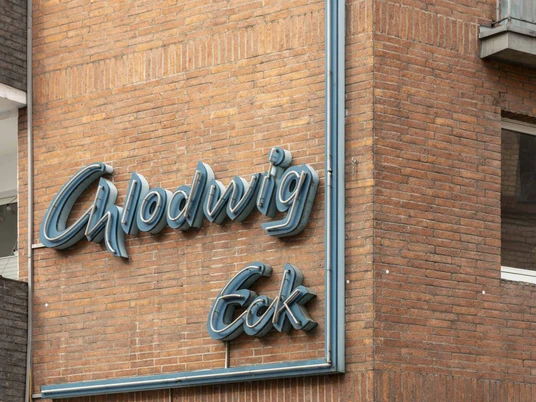 Chlodwig Eck Neonschriftzug "Chlodwig Eck" an einem Backsteingebäude, leicht verwittert, darunter das Eingangsschild.Neon lettering "Chlodwig Eck" on a brick building, slightly weathered, with the entrance sign underneath.