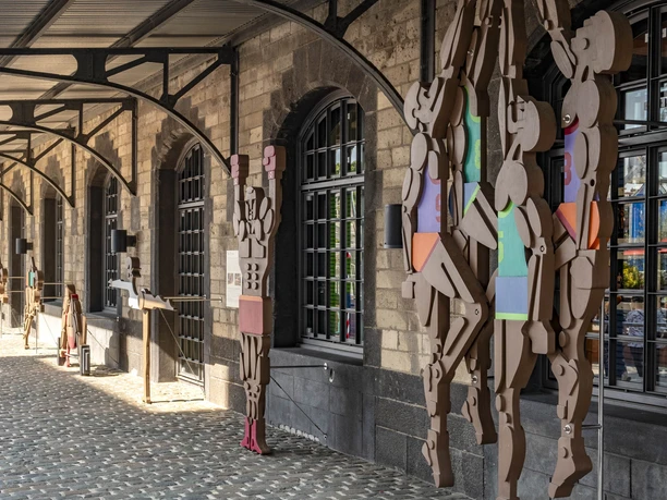 Deutsches Sport & Olympia Museum - Aussenansicht Das Bild zeigt eine überdachte Terrasse mit Figuren aus geometrischen Formen, die an Säulen lehnen.