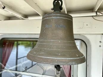 Die Schiffsglocke der MS "Stadt Verden" hängt vor einer Glasfläche mit rot-weißen Vorhängen.