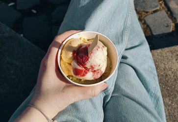 Madeleines-Homemade-Ice-Cream-Eisdiele-Bielefeld-Teutoburger-Wald-Tourismus.png Eine Hand hält einen Becher mit Vanille- und Erdbeereis, sitzend auf einer Pflastersteintreppe.