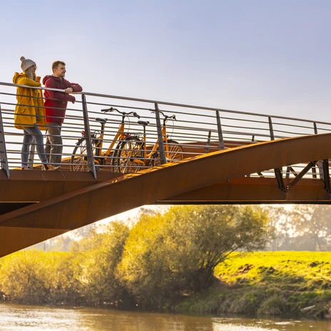 Fährbrücke über Ems zwischen Schwefingen und Klein Hesepe – Radfahren im Emsland ©Emsland Tourismus GmbH (2).jpg Zwei Radfahrende stehen auf einer modernen Brücke über der Ems und blicken in die Landschaft.