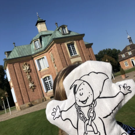 jpg (8).jpg Eine Person hält eine weiße Handpuppe mit Schlossmotiv vor einem historischen Backsteinbau.