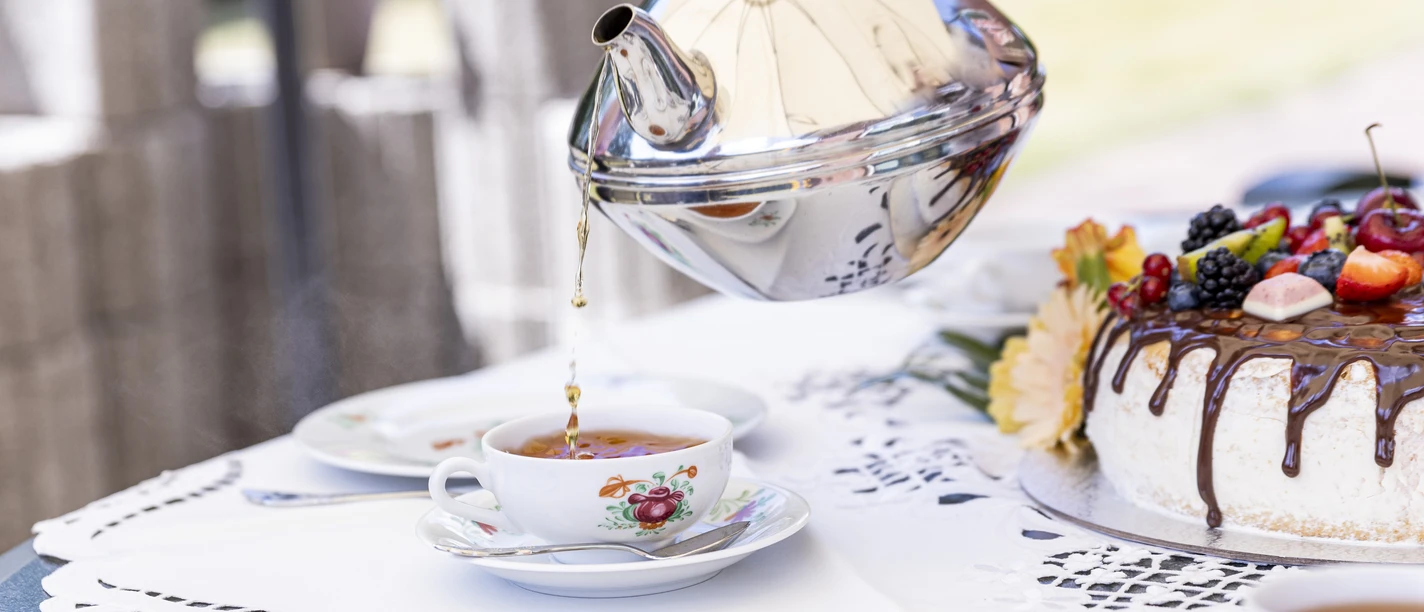 Teetied Thee wordt uit een elegante metalen pot in een porseleinen kopje geschonken, terwijl er een weelderige slagroomtaart met vers fruit naast staat.