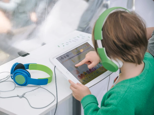 Hörspielwiese Cologne A child is sitting at a table, wearing green headphones and operating a tablet. Another pair of headphones is lying next to it.