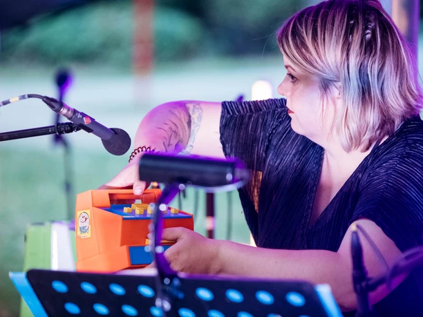 Hörspielwiese Köln Eine Frau mit Mikrofon hält ein orangefarbenes Spielzeug in der Hand, umgeben von Musik-Equipment.