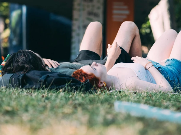 Two people lying on grass under shade at an open-air event with trees in the background