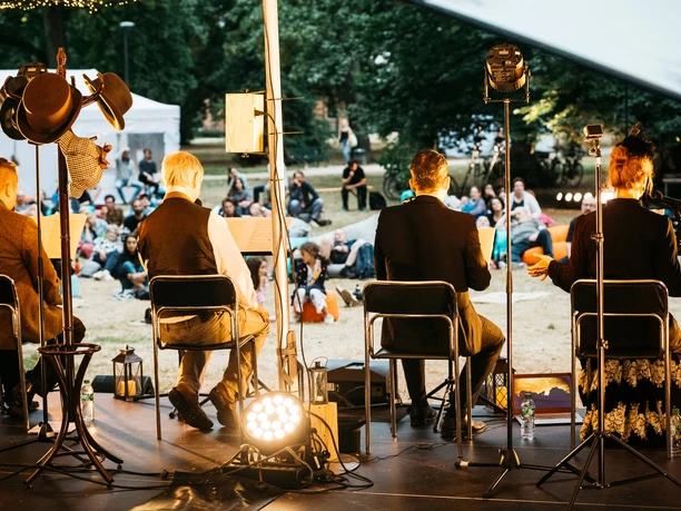 Hörspielwiese Cologne At the back of a stage, several older seated people are giving a reading to a large open-air audience.