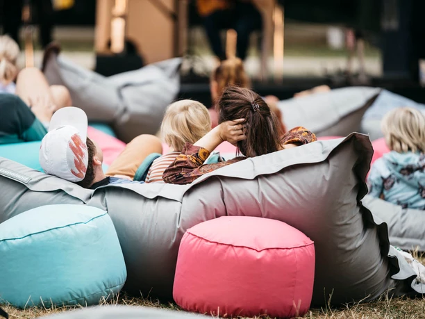 Hörspielwiese Cologne The picture shows people lying relaxed on colorful beanbags in the open air at the Hörspielwiese. A stage can be seen in the background, creating a relaxed and cozy atmosphere.
