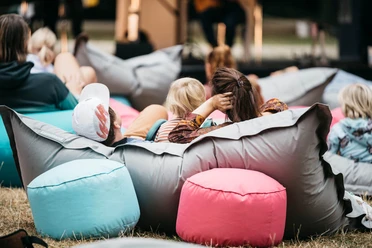 Hörspielwiese Köln Das Bild zeigt Menschen, die entspannt auf bunten Sitzsäcken im Freien bei der Hörspielwiese liegen. Eine Bühne ist im Hintergrund zu sehen, es herrscht eine ungezwungene und gemütliche Atmosphäre.