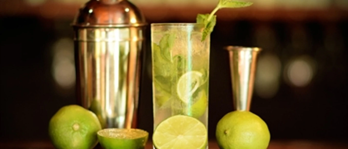 Cocktail - Workshop an der Bar Ein Mojito-Cocktail mit Limetten, Minze und einem Cocktailshaker auf einer Bar, sanft beleuchtet.