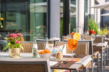 Taste & Cheers - Auf der Terrasse Aperitifs and snacks are served on white tablecloths on a sunny terrace with modern rattan furniture.