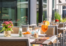 Taste & Cheers - Auf der Terrasse Aperitifs and snacks are served on white tablecloths on a sunny terrace with modern rattan furniture.