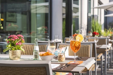 Taste & Cheers - Auf der Terrasse Aperitifs and snacks are served on white tablecloths on a sunny terrace with modern rattan furniture.