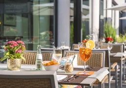 Taste & Cheers - Auf der Terrasse Auf einer sonnigen Terrasse mit modernen Rattanmöbeln stehen Aperitifs und Snacks auf weißen Tischtüchern.Aperitifs and snacks are served on white tablecloths on a sunny terrace with modern rattan furniture.