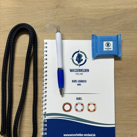 WasserheldenEmsland2.jpg Notizbuch mit Logo der Wasserhelden Emsland, daneben Kugelschreiber, Seil und verpackter Keks.