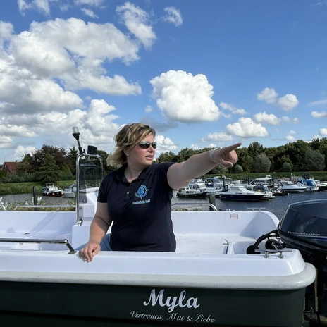 WasserheldenEmsland4.jpg Frau mit Sonnenbrille steuert ein Boot auf dem Wasser, zeigt Richtung Ufer mit Hafen und Booten.