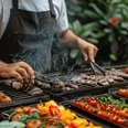 BBQ - Mit Live Cooking auf der Terrasse Grillmeister in schwarzer Schürze, der auf einem Barbecue-Grill verschiedene Fleischsorten und buntes Gemüse mit Grillzangen zubereitet, umgeben von frischen Zutaten und Rauchschwaden.Barbecue master in a black apron preparing various types of meat and colorful vegetables with barbecue tongs on a barbecue grill, surrounded by fresh ingredients and billows of smoke.