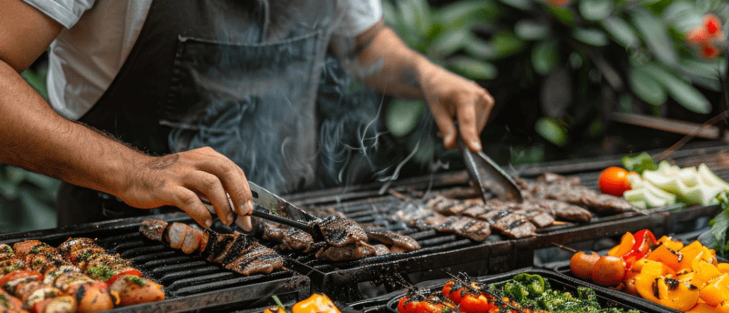 BBQ - Mit Live Cooking auf der Terrasse Barbecue master in a black apron preparing various types of meat and colorful vegetables with barbecue tongs on a barbecue grill, surrounded by fresh ingredients and billows of smoke.