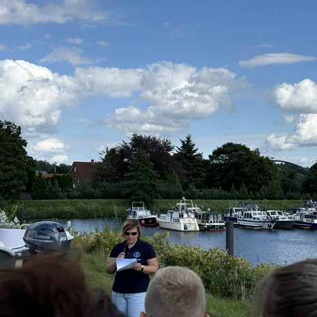 WasserheldenEmsland13.jpg Frau spricht am Ufer eines Kanals mit Booten im Hintergrund unter blauem Himmel mit Wolken.