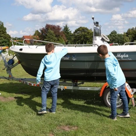 WasserheldenEmsland15.jpg Drei Kinder in blauen Pullovern spielen mit einem Boot auf einem Anhänger auf einer grünen Wiese.