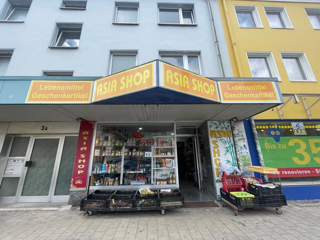 Asia Shop.jpg Zu sehen ist das Schaufenster und die Eingangstür des Asia Shops. Davor stehen Körbe mit frischen Lebensmitteln.
