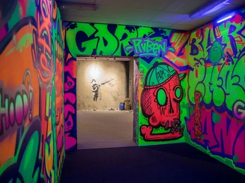 18 House of Banksy - Leipzig - Foto Andreas Schmidt.jpg Neonfarbene Wände mit Graffiti und im Hintergrund ein Banksy-Wandgemälde eines Mädchens.Neon-colored walls with graffiti and a Banksy mural of a girl in the background.Neonové zdi s graffiti a Banksyho nástěnnou malbou dívky v pozadí.Neonowe ściany z graffiti i muralem Banksy'ego przedstawiającym dziewczynę w tle.Neonkleurige muren met graffiti en een Banksy muurschildering van een meisje op de achtergrond.Pareti colorate al neon con graffiti e un murale di Banksy raffigurante una ragazza sullo sfondo.