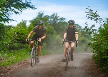 Zwei Gravelbiker fahren auf staubigem Waldweg durch dichtes Grün