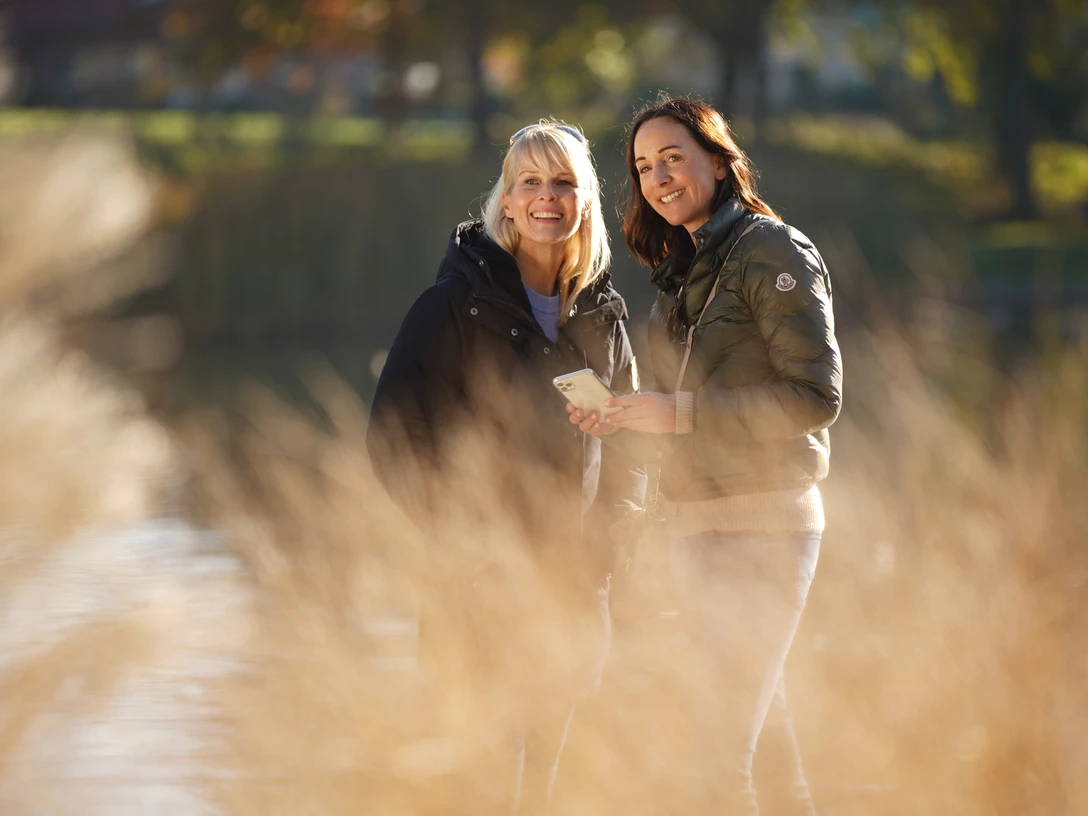 LM801971©tol-foto-lemon8.JPG Zwei Frauen stehen lächelnd in einem sonnigen Park vor verschwommenem Gras.