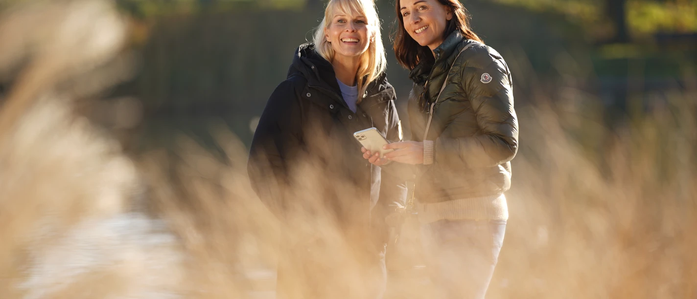 LM801971©tol-foto-lemon8.JPG Zwei Frauen stehen lächelnd in einem sonnigen Park vor verschwommenem Gras.