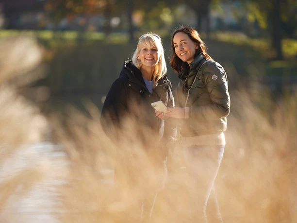 LM801971©tol-foto-lemon8.JPG Zwei Frauen stehen lächelnd in einem sonnigen Park vor verschwommenem Gras.