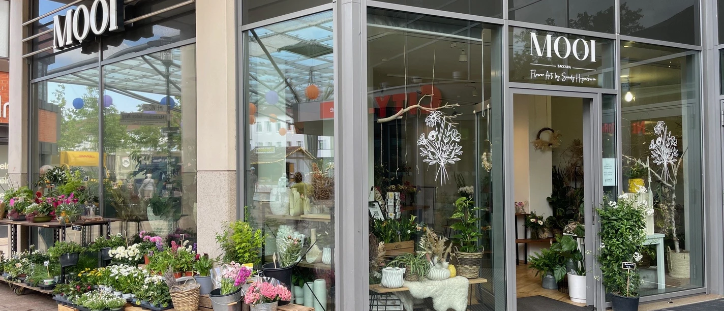 MOOI.jpg Zu sehen sind die Schaufenster und der Eingangsbereich des Blumengeschäfts. Vor den Schaufenstern sind verschiedene Blumen und Pflanzen ausgestellt.