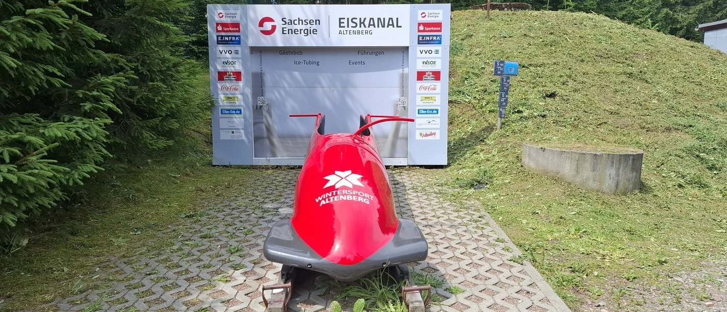 Fotopoint Bob SachsenEnergie-Eiskanal Altenberg.jpg Ein roter Bob vor der SachsenEnergie-Eiskanal Informationstafel in grüner Umgebung.