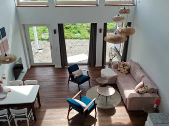 Finkennest.jpg Wohnzimmer mit hohen Decken und großen Fenstern, modern eingerichtet mit rosa Sofa und Esstisch.