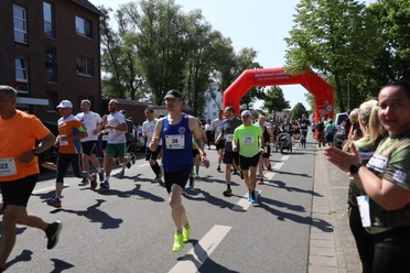 Stadtlauf Neustadt TSV Menschen unterschiedlichen Alters laufen konzentriert auf einer Straße unter einem roten Startbogen.