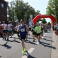 Stadtlauf Neustadt TSV Menschen unterschiedlichen Alters laufen konzentriert auf einer Straße unter einem roten Startbogen.