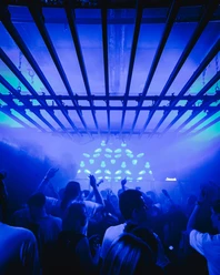 Bootshaus Dreherei Das Bild zeigt eine lebhafte Clubszene mit tanzenden Menschen unter blauer Beleuchtung und einem metallischen Gitter.
