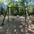 Der Spielplatz am Speukebusch zeigt eine hölzerne Schaukel vor einem Wald. Im Hintergrund sind weitere Spielgeräte.