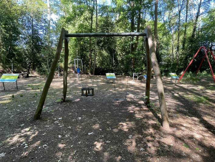 Kinderspielplatz Am Speukebusch Der Spielplatz am Speukebusch zeigt eine hölzerne Schaukel vor einem Wald. Im Hintergrund sind weitere Spielgeräte.