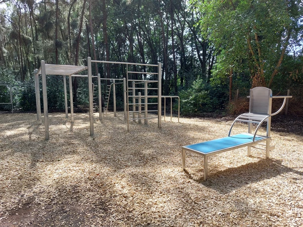 Links mehrteilige Klettergerüste aus Metall; rechts eine blaue Ruhebank. hölzerner Bodenbelag. Bäume umgeben den Platz.