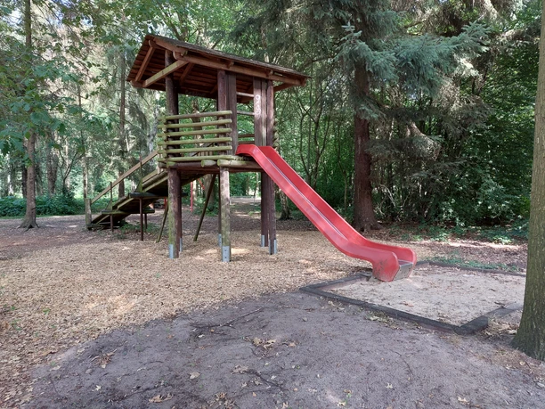 Kinderspielplatz Am Speukebusch Ein Spielturm aus Holz mit roter Rutsche steht im Wald. Der Boden ist mit Holzschnitzeln bedeckt.