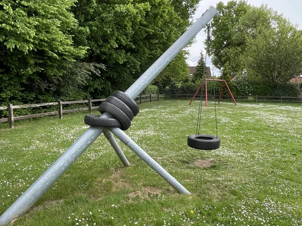 Spielplatz Auf dem Flage Klettergerüst aus großen Metallstangen mit alten Autoreifen auf grüner Wiese, umgeben von Bäumen.