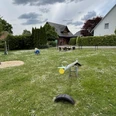 Ein Spielplatz mit Wippe, Rasenfläche und Sitzmöglichkeiten, umgeben von Häusern und Bäumen. Wolken am Himmel.