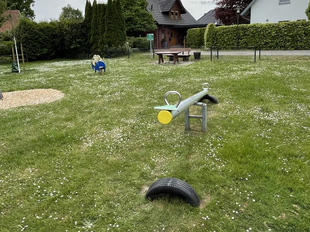 Ein Spielplatz mit Wippe, Rasenfläche und Sitzmöglichkeiten, umgeben von Häusern und Bäumen. Wolken am Himmel.