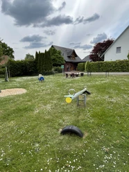 Spielplatz Auf dem Flage Ein Spielplatz mit Wippe, Rasenfläche und Sitzmöglichkeiten, umgeben von Häusern und Bäumen. Wolken am Himmel.