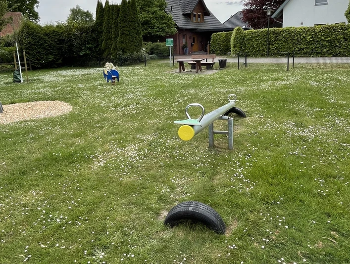 Spielplatz Auf dem Flage Ein Spielplatz mit Wippe, Rasenfläche und Sitzmöglichkeiten, umgeben von Häusern und Bäumen. Wolken am Himmel.