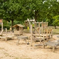 Ein großer Sandspielplatz mit vielfältigen Holzspielgeräten, Schaukeln, Kletterstrukturen und Bäumen.