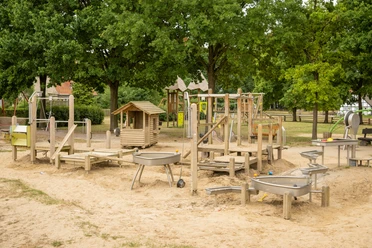 Spielplatz Museumshof Ein großer Sandspielplatz mit vielfältigen Holzspielgeräten, Schaukeln, Kletterstrukturen und Bäumen.