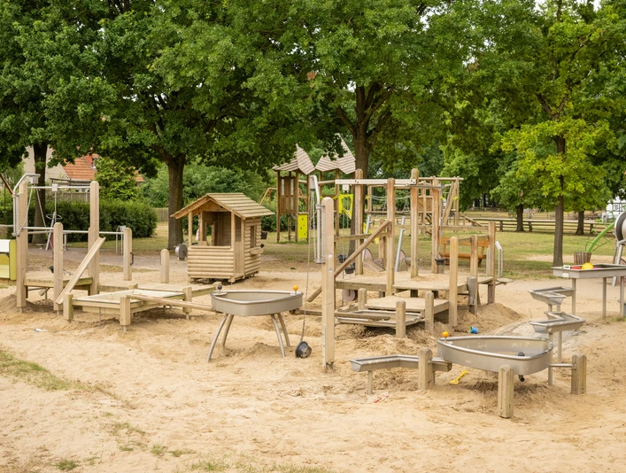 Spielplatz Museumshof Ein großer Sandspielplatz mit vielfältigen Holzspielgeräten, Schaukeln, Kletterstrukturen und Bäumen.