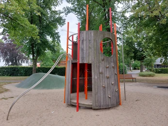 Spielplatz Alter Markt Das Bild zeigt einen modernen Spielplatz mit einem Holzturm und einer Rutsche. Drumherum sind grüne Bäume, und der Boden ist sandig. Links neben dem Turm befindet sich ein künstlicher Hügel. Die Umgebung wirkt freundlich und einladend für Kinder.