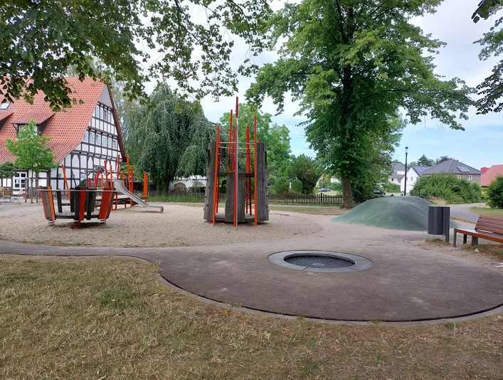 Ein moderner Spielplatz im Grünen mit Klettergerüst, Rutsche und Karussell. Ein Fachwerkhaus im Hintergrund.